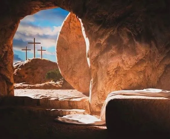View from inside an empty rock tomb with grave cloths lying on a stone ledge, looking out past the rolled-away stone to three crosses on a hill under a blue sky