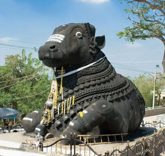 Giant black stone statue of a seated bull, decorated with flower garlands and white markings, on a raised platform behind a railing