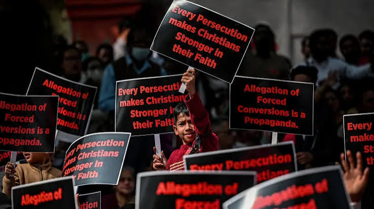 Christians at a protest holding black placards reading “Every Persecution makes Christians Stronger in their Faith” and “Allegation of Forced Conversions is a Propaganda,” with a young boy holding a sign up in the center