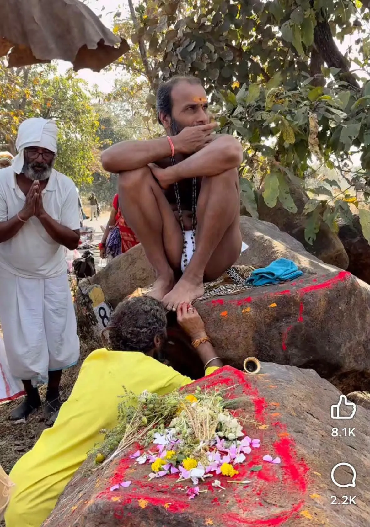 A person in yellow bows to touch the feet of a holy man squatting on a rock smeared with red powder and flowers, while an older man in white stands nearby with folded hands